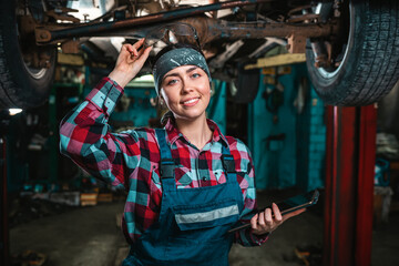 Obraz na płótnie Canvas Portrait of a young happy female mechanic in uniform posing with tablet at her hands and remove glasses from the head. The car is located on the lift