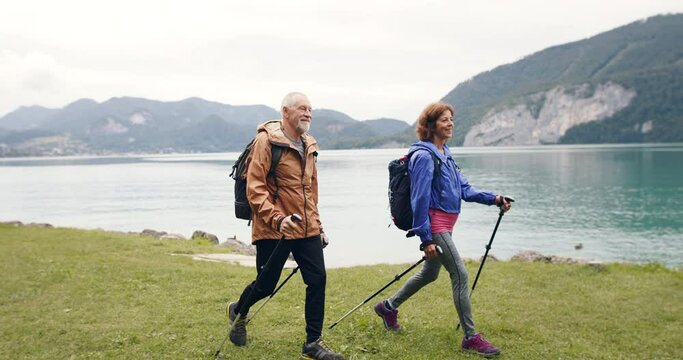 A side view of senior pensioner couple hiking by lake in nature.