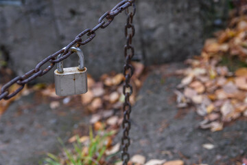 Rusty chain protects the entrance to the boat station. Old and worn chrome padlock hangs on the chain. Fallen leaves lie on a walkway. Property protection.