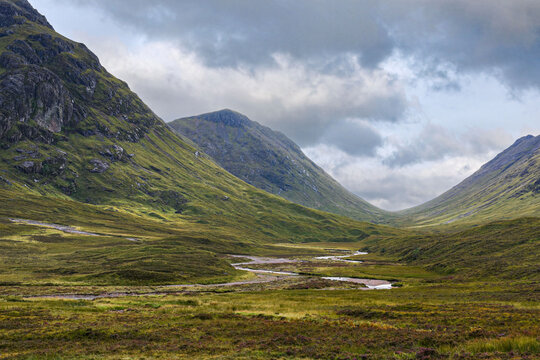 Glencoe valley in the Highlands of Scotland. The Glencoe river meanders between rocky mountain slopes across moorland covered with wild flowers and purple heather.