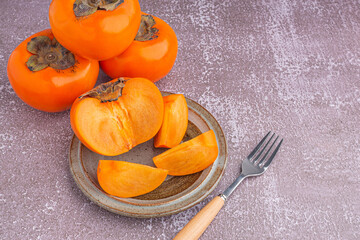 Fresh ripe persimmons with slices of persimmon on a plate with cutlery and whole fruit on gray stone background. Space for text. Close-up photo. Concept of health fruit