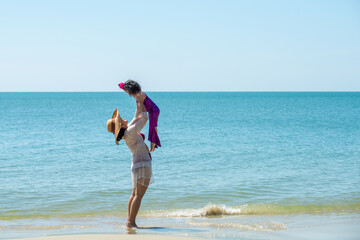 Happy Asian family holiday vacation. Beautiful Asian mother play and carry little daughter up in the air on the beach in summer day. Cute child girl and mom relax and having fun together on the beach