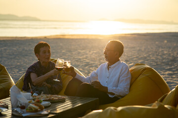Happy Asian family on summer holiday vacation. Healthy senior couple sit on the beach and enjoy romantic anniversary dinner party with drinking wine. Retired man and woman relaxing weekend activity