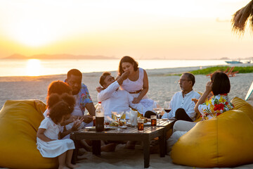 Group of multiethnic family friends enjoy dinner party together on the beach at sunset. Diverse...