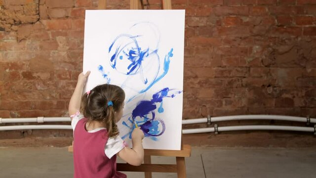 Pretty little three year old girl draws with blue paint on canvas on an easel, finishing up her work, adding little details to the painting. Children's creativity. Close-up shot of painting process. 
