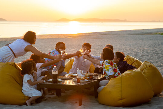 Group Of Multiethnic Family Friends Enjoy Dinner Party Together On The Beach At Sunset. Diverse Family With Child Girl, Adult And Senior Couple Relax And Having Fun Together On Summer Holiday Vacation