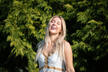 A beautiful young woman with long curly hair wearing a dress poses near trees