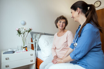 Fototapeta premium Senior Woman and Home Nurse Sitting on Bed Smiling at Each Other. Cheerful Elderly Female Patient Looking At Home Caregiver in Hospital Room.