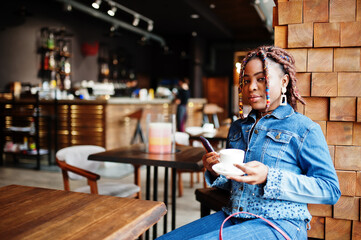 Lovely african american woman with dreadlocks in blue stylish jeans jacket at cafe hold mobile phone. Beautiful cool fashionable black young girl indoor drink coffee.