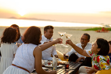 Group of multiethnic family friends enjoy dinner party together on the beach at sunset. Diverse family with child girl, adult and senior couple relax and having fun together on summer holiday vacation