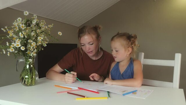 Mother and daughter draw with colored pencils in large album home. Scandinavian family blonde girls with freckles paints children's drawings. Girls are sitting white table in room with Daisy flowers