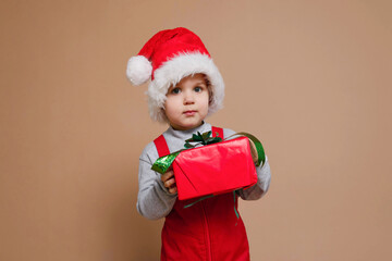 Funny toddler boy holds a Christmas gift in isolation against the background of the trendy color Set Sail Champagne. Holidays, traditions concept.