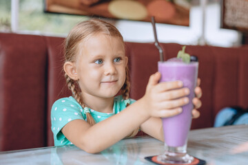 A little cute child with blond pigtails is looking at a large glass of milkshake and enjoying a sweet dessert in a local cozy cafe. Portrait.