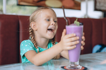 A little adorable child with blond pigtails is looking at a large glass of milkshake and enjoying a sweet dessert in a local cozy cafe. Portrait.
