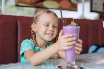 A little beautiful girl with blond pigtails is looking at a large glass of milkshake and enjoying a sweet dessert in a local cozy cafe. Portrait.