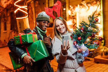 Close up of joyful African American man with Caucasian woman on street while taking selfie photo on smartphone. Happy multi-ethnic couple standing outdoors and taking pictures together with xmas gift