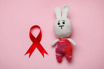 Cute knitted toy bunny and red ribbon on pink background, flat lay. AIDS disease awareness