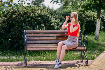 A woman sits on a park bench in a skirt and a red top and puts on sunglasses.