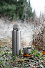 Vacuum thermos with a mug in the winter forest. Steel travel flask and a glass of steam. Concept for hot winter drink, travel, hike. Vertical photo