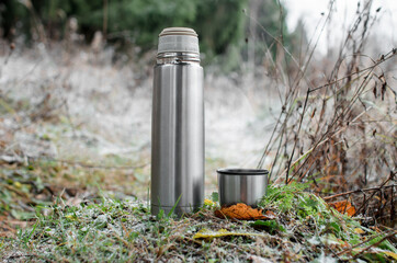 Vacuum thermos with a mug in the winter forest. Steel travel flask and a glass of steam. Concept for hot winter drink, travel, hike.