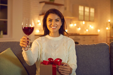 Happy woman holding glass of wine and holiday present during online Valentines day celebrating