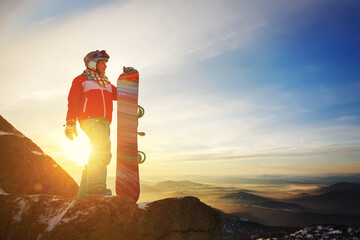 Girl snowboarder stands with snowboard on mountain's top on sunset backdrop.