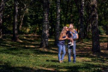 Mom kisses her daughter sitting in her father's arms. The family is resting in nature. Summer sunny day. Copy space.