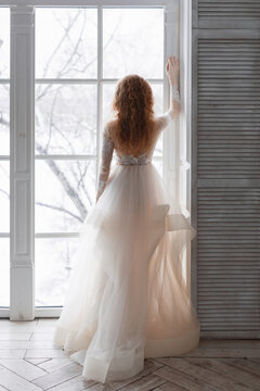 Unrecognizable slim pretty girl in white wedding luxurious dress stands in front of the window. Ginger curly bride in floor-length chic wedding dress waiting a groom. Vertical photo. Marriage concept.