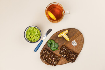 Wooden board with ingredients for avocado toast. Dark rye bread with avocado slices, fresh basil leaves, lemon slices and sesame seeds. Near guacamole in bowl and cup of tea. Healthy breakfast concept
