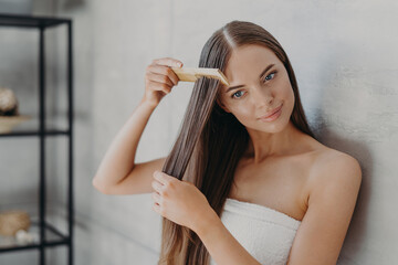 Young brunette woman brushes hair with comb after taking shower and applying hair care mask, wears minimal makeup, has healthy glowing skin after hygienic procedures, stands wrapped in towel - Powered by Adobe