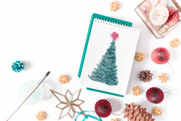 Christmas composition. An open notebook with a watercolor painted Christmas tree is on a white background. There are Christmas decorations and a paintbrush nearby. Top view, flat lay, copy space.