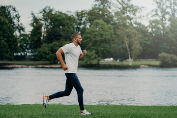 Athlete bearded man runs along beautiful landscape, demonstrates endurance, dressed in active wear,...