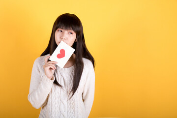 Portrait of lovely shy asian girl in white sweater holding valentine's day greeting card and missing someone. Valentine's day concept photos. Studio shot isolated on yellow background.