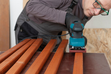 Carpenter working with electric sheet finishing sander on wooden  slats for production of  wooden garden bench on table in carpentry  workshop