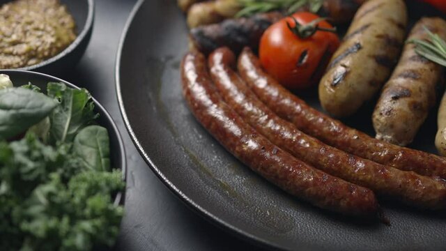 The cook with tongs takes the grilled sausage from a plate with assorted kobasoks, lettuce, vegetables, mustard. The camera moves forward, close-up