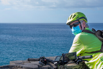 Coronavirus. Portrait of a senior man with bicycle and helmet in sport activity enjoying nature and sea, wearing a protective face mask due to coronavirus contagion