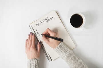 Female hands writing My Goals 2021 in a notebook. Mug of coffee on the table, top view