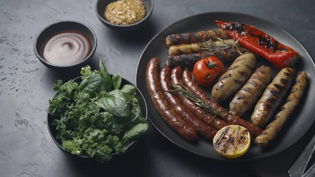 Plate with grilled sausages, vegetables, tomato sauce, mustard and lettuce on a dark background, close-up, the camera moves to the side, place for text