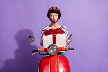 Portrait photo of shocked girl sitting on scooter keeping big present box opened mouth helmet isolated on bright purple color background