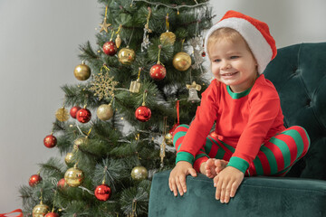 excited toddler blonde baby girl in Santa hat sitting on chair near the Christmas tree.