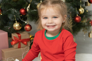 close up portrait excited toddler blonde baby girl in red jacket sitting near the Christmas tree.
