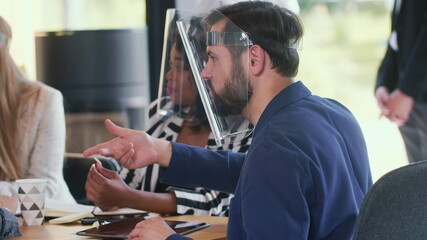 Side view serious young male Caucasian boss leads business team at office meeting, all wear COVID-19 safety face shields