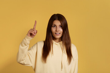 Im genius. Serious caucasian woman in yellow hoodie has enthusiastic expression raised index finger, happy to find solution. Isolated over a yellow background.