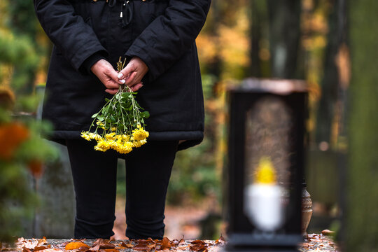 Mourning woman holding flowers in hands and standing at grave in cemetery. Sadness during funeral