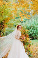 Obraz na płótnie Canvas Sweet red-haired bride stands in autumn park with a bouquet of flowers in her hands.
