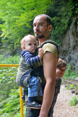 Naklejka na ściany i meble Travel with small children. Father with baby in carrier and ten year old son walking in mountain gorge.