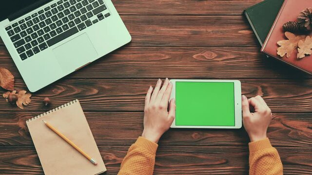 Top view of girl in yellow sweater sitting at wooden table and reading article online on digital tablet with green screen.