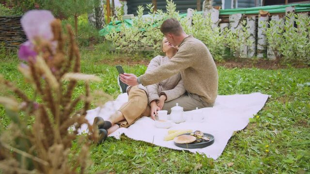 A man and a woman on a picnic in the garden take a selfie on a smartphone and kiss in slowmotion