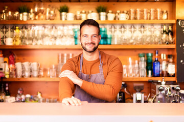 Small business owner man standing in the restaurant and waiting for guest. Portrait shot of handsome waiter wearing apron and smiling while standing behind the counter in the cafe