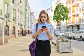 Fototapeta premium Young business woman with smartphone walking along city street